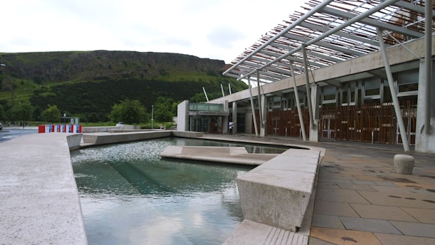 A modern architectural structure with a water feature in the foreground. The building has a unique design with an overhanging metal framework and large glass windows. The background features a lush green hillside, adding a natural contrast to the urban setting.