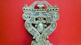 An intricately carved wooden object with floral patterns, ornate scrolls, and symmetrical design against a solid red background. The craftsmanship displays a vintage and artistic feel, with attention to detail in the carvings.