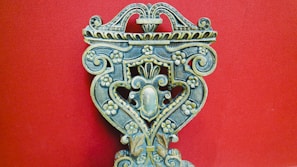An intricately carved wooden object with floral patterns, ornate scrolls, and symmetrical design against a solid red background. The craftsmanship displays a vintage and artistic feel, with attention to detail in the carvings.