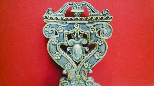 An intricately carved wooden object with floral patterns, ornate scrolls, and symmetrical design against a solid red background. The craftsmanship displays a vintage and artistic feel, with attention to detail in the carvings.
