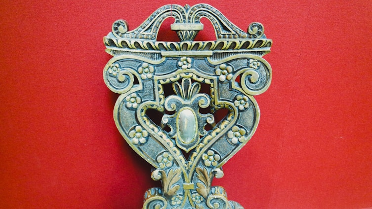 An intricately carved wooden object with floral patterns, ornate scrolls, and symmetrical design against a solid red background. The craftsmanship displays a vintage and artistic feel, with attention to detail in the carvings.
