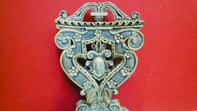 An intricately carved wooden object with floral patterns, ornate scrolls, and symmetrical design against a solid red background. The craftsmanship displays a vintage and artistic feel, with attention to detail in the carvings.