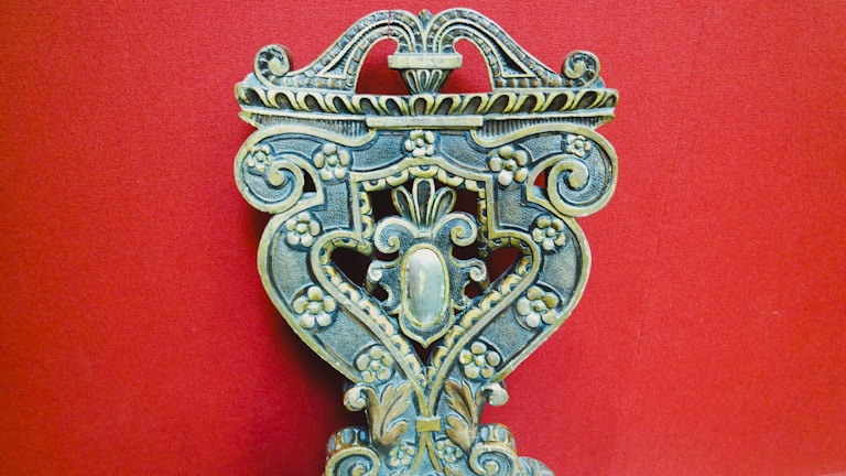 An intricately carved wooden object with floral patterns, ornate scrolls, and symmetrical design against a solid red background. The craftsmanship displays a vintage and artistic feel, with attention to detail in the carvings.