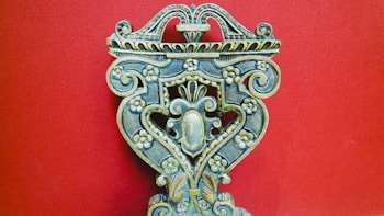 An intricately carved wooden object with floral patterns, ornate scrolls, and symmetrical design against a solid red background. The craftsmanship displays a vintage and artistic feel, with attention to detail in the carvings.