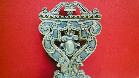 An intricately carved wooden object with floral patterns, ornate scrolls, and symmetrical design against a solid red background. The craftsmanship displays a vintage and artistic feel, with attention to detail in the carvings.