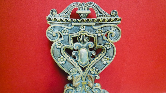 An intricately carved wooden object with floral patterns, ornate scrolls, and symmetrical design against a solid red background. The craftsmanship displays a vintage and artistic feel, with attention to detail in the carvings.