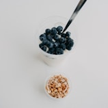 A glass filled with creamy yogurt topped with fresh blueberries is placed next to a small container of granola. A metallic spoon is immersed in the yogurt. The background is plain white, creating a clean and minimalist aesthetic.