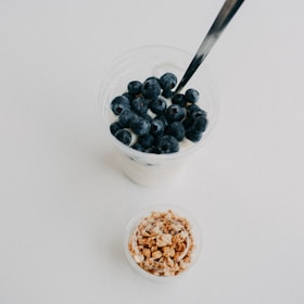 A glass filled with creamy yogurt topped with fresh blueberries is placed next to a small container of granola. A metallic spoon is immersed in the yogurt. The background is plain white, creating a clean and minimalist aesthetic.