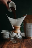 A hand pours coffee grounds from a small cup into a glass coffee dripper, which sits on a wooden surface. A paper filter is placed inside the dripper. In the background, there is a ceramic mug with a geometric pattern and a package labeled 'Community Blend.' The setting appears cozy and rustic, suggesting a calm and inviting environment.