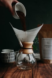 A hand pours coffee grounds from a small cup into a glass coffee dripper, which sits on a wooden surface. A paper filter is placed inside the dripper. In the background, there is a ceramic mug with a geometric pattern and a package labeled 'Community Blend.' The setting appears cozy and rustic, suggesting a calm and inviting environment.