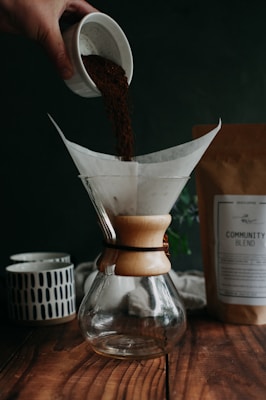 A hand pours coffee grounds from a small cup into a glass coffee dripper, which sits on a wooden surface. A paper filter is placed inside the dripper. In the background, there is a ceramic mug with a geometric pattern and a package labeled 'Community Blend.' The setting appears cozy and rustic, suggesting a calm and inviting environment.