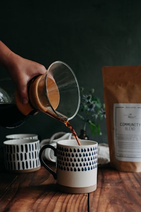 A hand pours coffee from a glass carafe into a ceramic mug with a blue drop pattern on a wooden table. Other similar mugs and a brown paper coffee bag labeled 'Community Blend' are also on the table. The background is dark with a soft focus on a plant, lending a cozy, rustic ambiance.