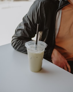 A plastic cup filled with an iced beverage sits on a white table. The drink appears to be creamy with ice cubes visible, and it has a black straw inserted. Part of a person in a black jacket and peach-colored shirt is visible, with an arm resting on the table.