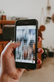A hand is holding a smartphone displaying an Instagram post with the hashtag '#workspacegoals' visible on the screen. The background features a blurred image of a cozy room with wooden furniture and plants.