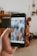 A hand is holding a smartphone displaying an Instagram post with the hashtag '#workspacegoals' visible on the screen. The background features a blurred image of a cozy room with wooden furniture and plants.