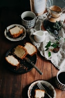 Slices of cake are arranged on rustic plates and a black tray, surrounded by cups of coffee, a Chemex coffee maker, a white pitcher, and a small vase with green leaves and flowers on a wooden table. A bag of coffee beans and a linen cloth add a cozy touch.