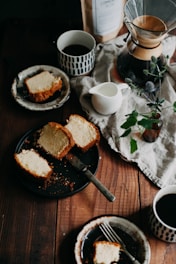 Slices of cake are arranged on rustic plates and a black tray, surrounded by cups of coffee, a Chemex coffee maker, a white pitcher, and a small vase with green leaves and flowers on a wooden table. A bag of coffee beans and a linen cloth add a cozy touch.