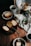 Slices of cake are arranged on rustic plates and a black tray, surrounded by cups of coffee, a Chemex coffee maker, a white pitcher, and a small vase with green leaves and flowers on a wooden table. A bag of coffee beans and a linen cloth add a cozy touch.