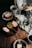 Slices of cake are arranged on rustic plates and a black tray, surrounded by cups of coffee, a Chemex coffee maker, a white pitcher, and a small vase with green leaves and flowers on a wooden table. A bag of coffee beans and a linen cloth add a cozy touch.