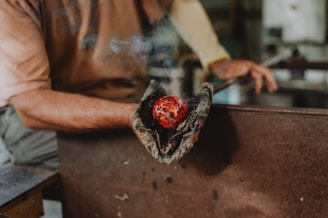 A person is holding a hot, glass-blown object with protective gloves. The object is red and has a marbled appearance. There is some smoke emanating from the gloves, indicating the high temperature of the glass.