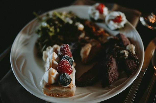 A white plate contains a variety of foods including a dessert topped with whipped cream, raspberries, and blueberries, slices of cooked meat, and a green salad. Smaller bites with cream and red garnish are placed at the top of the plate.