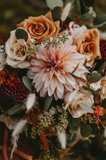 A beautifully arranged bouquet featuring a large pink dahlia at the center, surrounded by brown and white roses, greenery, and dried elements that add texture. The composition of the flowers is artful and balanced, creating a rustic and natural aesthetic.