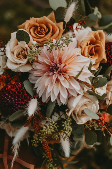 A beautifully arranged bouquet featuring a large pink dahlia at the center, surrounded by brown and white roses, greenery, and dried elements that add texture. The composition of the flowers is artful and balanced, creating a rustic and natural aesthetic.