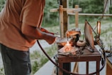 A person is working with a small torch on a wooden table, which is cluttered with various tools and metal scraps. A small flame is visible, and there's a fabric-covered object nearby. The background is an outdoor setting with greenery visible.
