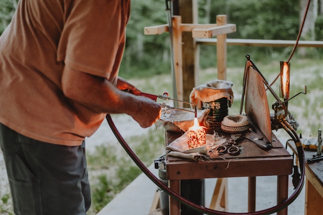 A person is working with a small torch on a wooden table, which is cluttered with various tools and metal scraps. A small flame is visible, and there's a fabric-covered object nearby. The background is an outdoor setting with greenery visible.
