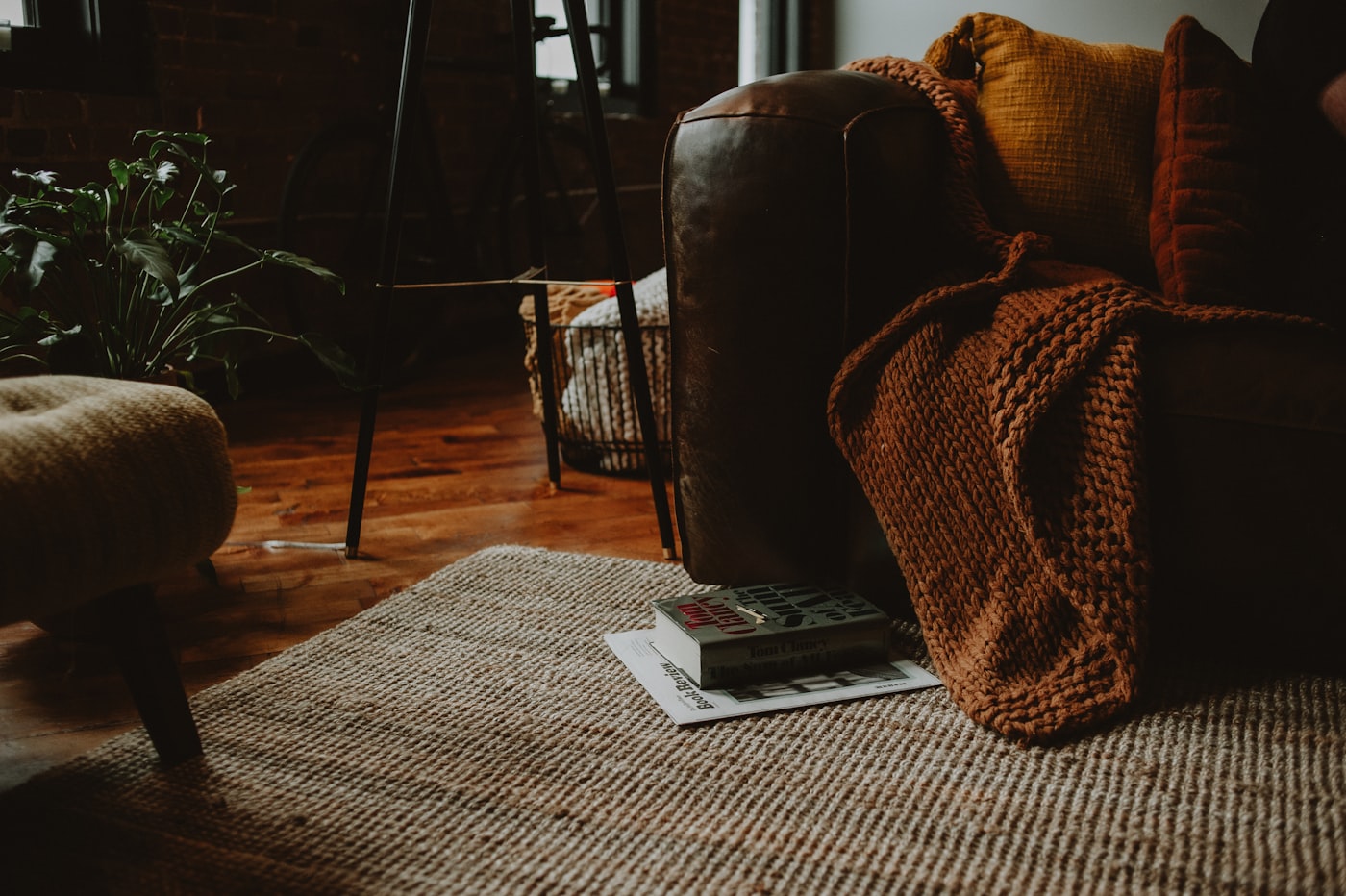 Cozy apartment living room corner with rich brown leather sofa, chunky rust-orange knit throw, jute woven rug, warm wood floors, and greenery in a brick-walled space