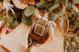 A perfume bottle labeled 'Victoria's Secret Bombshell' is surrounded by green leaves and beige high-heeled shoes, placed on a textured surface. The composition includes soft floral elements that add elegance and warmth.