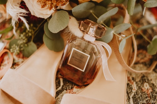 A perfume bottle labeled 'Victoria's Secret Bombshell' is surrounded by green leaves and beige high-heeled shoes, placed on a textured surface. The composition includes soft floral elements that add elegance and warmth.