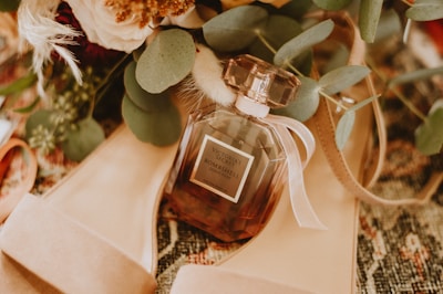 A perfume bottle labeled 'Victoria's Secret Bombshell' is surrounded by green leaves and beige high-heeled shoes, placed on a textured surface. The composition includes soft floral elements that add elegance and warmth.