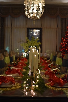 A beautifully decorated dining table set for a festive occasion. Tall white candles are placed at the center, surrounded by greenery and red ornamental napkins on white plates. The setting is complemented by a chandelier hanging from the ceiling, adding an elegant touch. A Christmas tree with red lights is visible in the background.