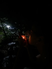 Shadowy figure lighting a match near a building engulfed in flames at dusk.
