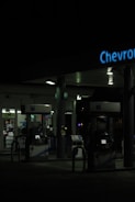 A dimly lit gas station at night featuring several gas pumps under a canopy with the logo partially visible. The station is mostly dark, with some interior lighting emanating from the convenience store in the background.
