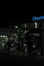 A dimly lit gas station at night featuring several gas pumps under a canopy with the logo partially visible. The station is mostly dark, with some interior lighting emanating from the convenience store in the background.