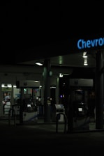 A dimly lit gas station at night featuring several gas pumps under a canopy with the logo partially visible. The station is mostly dark, with some interior lighting emanating from the convenience store in the background.