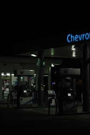 A dimly lit gas station at night featuring several gas pumps under a canopy with the logo partially visible. The station is mostly dark, with some interior lighting emanating from the convenience store in the background.