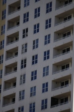 A tall residential apartment building with multiple floors and uniform balconies. The facade is a light color, with dark-framed windows evenly distributed across the structure. Each apartment appears to have large, floor-to-ceiling windows and minimalistic railings.