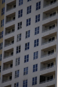 A tall residential apartment building with multiple floors and uniform balconies. The facade is a light color, with dark-framed windows evenly distributed across the structure. Each apartment appears to have large, floor-to-ceiling windows and minimalistic railings.