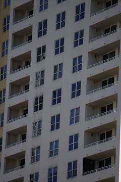 A tall residential apartment building with multiple floors and uniform balconies. The facade is a light color, with dark-framed windows evenly distributed across the structure. Each apartment appears to have large, floor-to-ceiling windows and minimalistic railings.