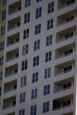 A tall residential apartment building with multiple floors and uniform balconies. The facade is a light color, with dark-framed windows evenly distributed across the structure. Each apartment appears to have large, floor-to-ceiling windows and minimalistic railings.