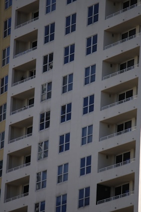 A tall residential apartment building with multiple floors and uniform balconies. The facade is a light color, with dark-framed windows evenly distributed across the structure. Each apartment appears to have large, floor-to-ceiling windows and minimalistic railings.