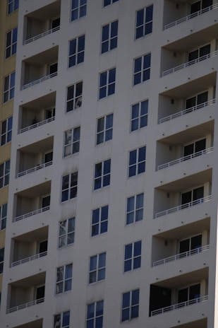 A tall residential apartment building with multiple floors and uniform balconies. The facade is a light color, with dark-framed windows evenly distributed across the structure. Each apartment appears to have large, floor-to-ceiling windows and minimalistic railings.