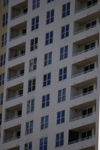 A tall residential apartment building with multiple floors and uniform balconies. The facade is a light color, with dark-framed windows evenly distributed across the structure. Each apartment appears to have large, floor-to-ceiling windows and minimalistic railings.