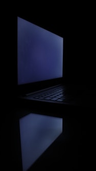 A dimly lit laptop screen glowing faintly in a dark environment, creating an atmospheric reflection on a smooth surface.