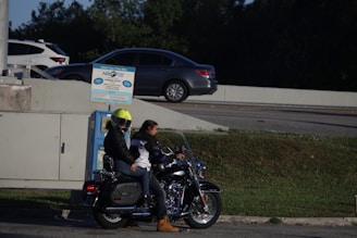 Two people are on a motorcycle parked near a wall. One person is wearing a bright yellow helmet and a black jacket, while the other is wearing a white and black jacket. Behind them, there is a tire inflation machine. Several cars are visible on a road in the background, and there are trees surrounding the area.