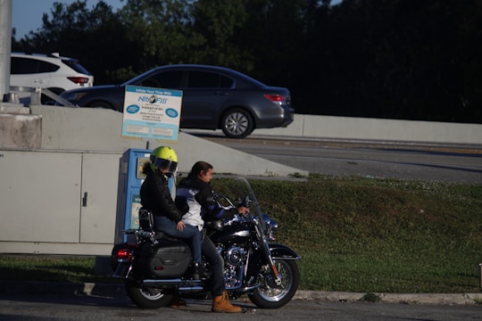 Two people are on a motorcycle parked near a wall. One person is wearing a bright yellow helmet and a black jacket, while the other is wearing a white and black jacket. Behind them, there is a tire inflation machine. Several cars are visible on a road in the background, and there are trees surrounding the area.