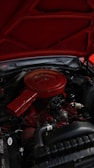 An engine compartment of a vehicle with a predominantly red air filter cover and various mechanical components. The hood is open, revealing parts like hoses, wires, and metallic elements surrounding the engine block.